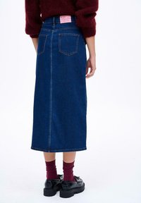 Denim maxi sukňa v stredne modrej farbe, s rovnou siluetou, zadnými vreckami a viditeľným centrálnym švom. Skombinovaná s čiernymi platformovými topánkami a vínovými ponožkami.