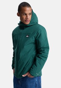 Groene geïsoleerde jas met een capuchon, voorzien van een ritssluiting, elastische manchetten en een klein logo op de borst, gecombineerd met jeans.