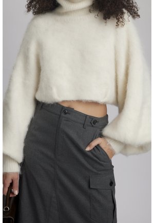 Femme portant un pull court en maille blanche duveteuse à col roulé et une jupe taille haute gris foncé avec poche cargo, main dans la poche.