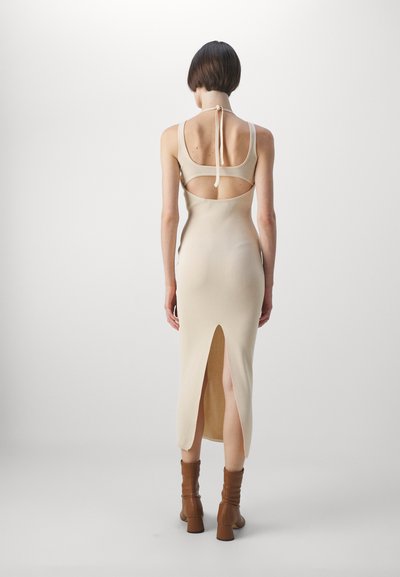 Beige, knee-length dress with cut-out back, thin straps, and a back tie. Features a side slit and soft, stretchy knit fabric. Brown heeled boots.