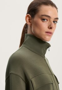 Olivno zelena jakna s visokim ovratnikom, ki ima zadrgo, subtilno teksturo in izrazite šive. Vidna sta dva sprednja žepa.