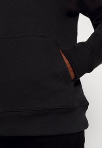 Main à la peau foncée partiellement à l'intérieur de la poche avant d'un sweat-shirt noir avec poignets et ourlet côtelés.