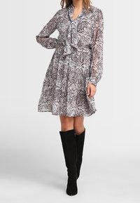 Woman wearing a patterned, long-sleeve, knee-length dress paired with black knee-high boots, standing with one hand on her hip.