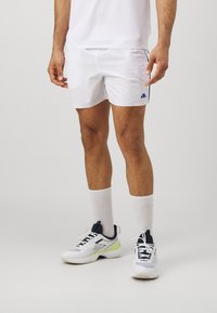 White athletic shorts with side pockets and blue accent logo, paired with white socks and white/navy sport shoes with green accents.
