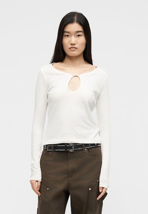 Jeune femme aux longs cheveux bruns portant un haut blanc à manches longues avec une découpe frontale et un pantalon marron avec une ceinture noire cloutée.