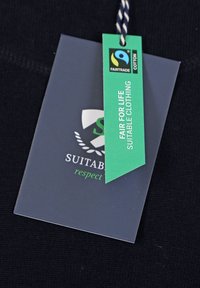 Zwarte stoffen label met groene Fairtrade-certificeringslabel, met de teksten "FAIR FOR LIFE" en "GESCHIKTE KLEDING", een gladde textuur en nette stiksel.