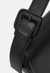 Gros plan sur la lanière en cuir noir d’un sac avec une boucle rectangulaire rembourrée et des bords cousus sur un fond uni.