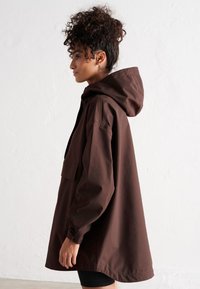 Chaqueta marrón con capucha, hecha de un tejido suave y ligero. Corte holgado con bolsillo frontal, mangas largas y dobladillo curvado. Diseño sencillo.
