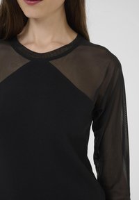 Mujer vistiendo una blusa negra con un cuerpo opaco y mangas largas de malla transparente en la parte superior, labios pintados con lápiz labial rosa oscuro.