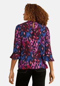 Krāsains blūzes tops ar purpursarkanu, zilu un sarkanu leopardu rakstu. Tam ir piegriezums ar trim ceturtdaļām, kurām ir viļņotas malas un atvērums aizmugurē.