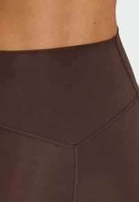 Legging marron taille haute avec une texture lisse, des coutures contrastées et un détail géométrique sur la ceinture.