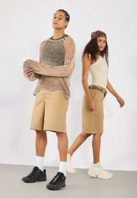 Deux mannequins portant des tenues décontractées. L'un porte un gros pull en maille beige et un short beige, l'autre porte un débardeur beige ajusté et une ceinture à motifs.