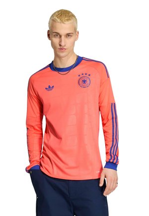 Jeune homme aux cheveux blonds courts, portant un maillot de sport coral à manches longues Adidas avec des rayures bleues et un pantalon bleu marine, debout avec la main dans la poche.