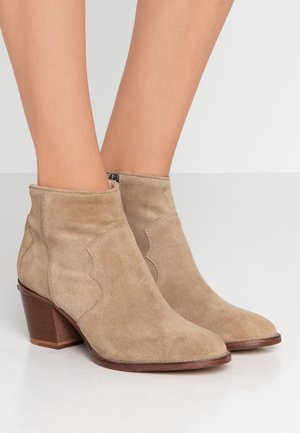 Bottines en daim beige à petit talon en bois empilé, portées sur jambes nues, avec fermeture éclair latérale et coutures décoratives subtiles.