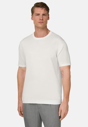 Mann steht und trägt ein weißes Kurzarm-T-Shirt mit Rundhalsausschnitt und hellgraue Hose vor neutralem Hintergrund.