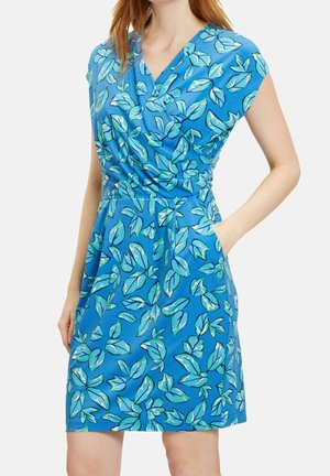 Kobieta w niebieskiej sukience bez rękawów z zielonym i białym nadrukiem liści, z kopertowym górnym krojem i bocznymi kieszeniami.