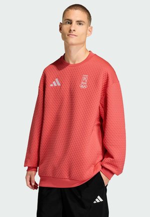 Młody mężczyzna z krótkimi włosami, ubrany w czerwoną, teksturowaną bluzę Team Olympijskiego i czarne spodnie Adidas, stoi z jedną ręką w kieszeni.