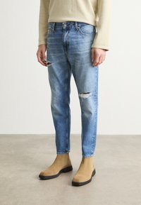Ljust blå denimjeans med rak passform, utrustade med slitna rippar vid knäna, bärs tillsammans med beige mocka chelsea-boots.