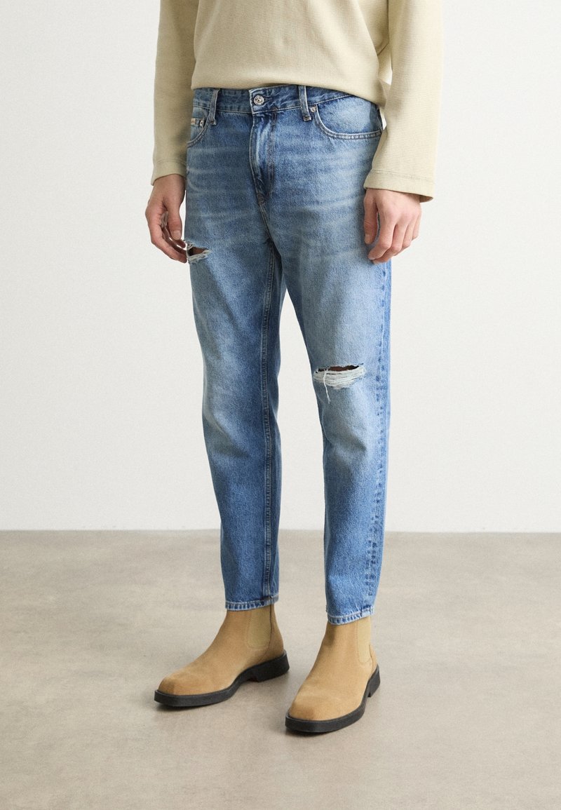 Ljust blå denimjeans med rak passform, utrustade med slitna rippar vid knäna, bärs tillsammans med beige mocka chelsea-boots.