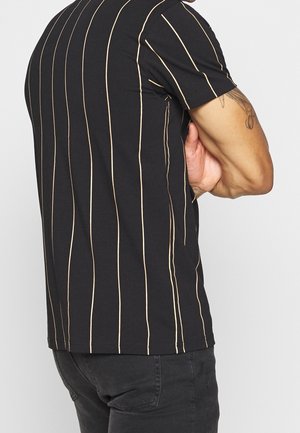 Hombre con camiseta negra de manga corta con finas rayas verticales beige, brazos cruzados, combinado con vaqueros oscuros, tatuaje visible en el brazo izquierdo.