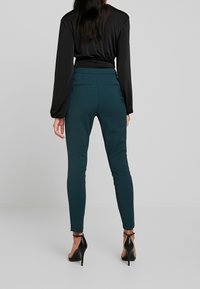 Svart långärmad topp som kombineras med figurnära turkosa byxor, med två bakfickor, och bärs med svarta högklackade skor.