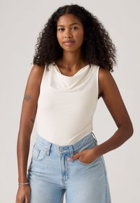 Frau mit lockigem Haar, die ein ärmelloses weißes Oberteil und hellblaue Jeans trägt, mit einer Hand in der Tasche und einem Tattoo am linken Arm.