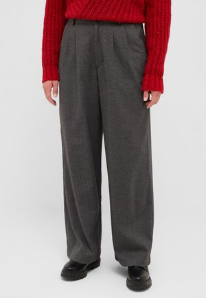 Graue weite Hose mit einem strukturierten Muster, ausgestattet mit vorderen Falten und seitlichen Taschen, kombiniert mit schwarzen Stiefeln und einem roten Strickpullover.