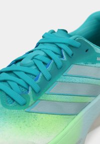 Τουρκουάζ αθλητικό παπούτσι με υφασμάτινη υφή, κορδόνια στον ίδιο τόνο και τρεις μεταλλικές ασημί ρίγες στο πλάι. Απαλή βαθμίδωση από τουρκουάζ σε πράσινο.