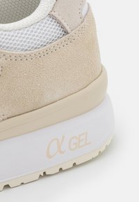Beige suede sneaker with mesh accents, featuring a white midsole with alpha gel branding, textured surface, and rounded heel design.