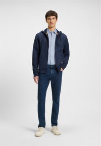 Sudadera con capucha azul marino con acentos naranjas, combinada con una camisa de botones azul claro y jeans de denim oscuro. El calzado presenta zapatillas de color crema.