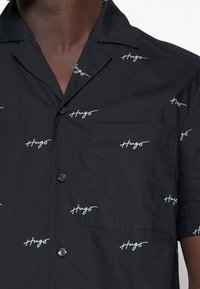 Korte mouwen, zwart shirt met een herhalend witte "Hugo" logo print. Voorkant met een linkerzak en knoopsluiting met een gesneden kraag.