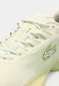 Αθλητικό παπούτσι Lacoste με εκρού επάνω μέρος, πάνελ από δίχτυ, πράσινες λάιμ λεπτομέρειες και το χαρακτηριστικό λογότυπο. Διαθέτει επίπεδα κορδόνια και υφή επιφάνειας.