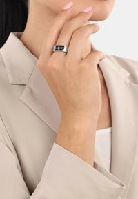 Anello in argento con finitura lucida, caratterizzato da accenti neri e motivi a strisce orizzontali, indossato su una mano con pelle chiara e unghie curate.