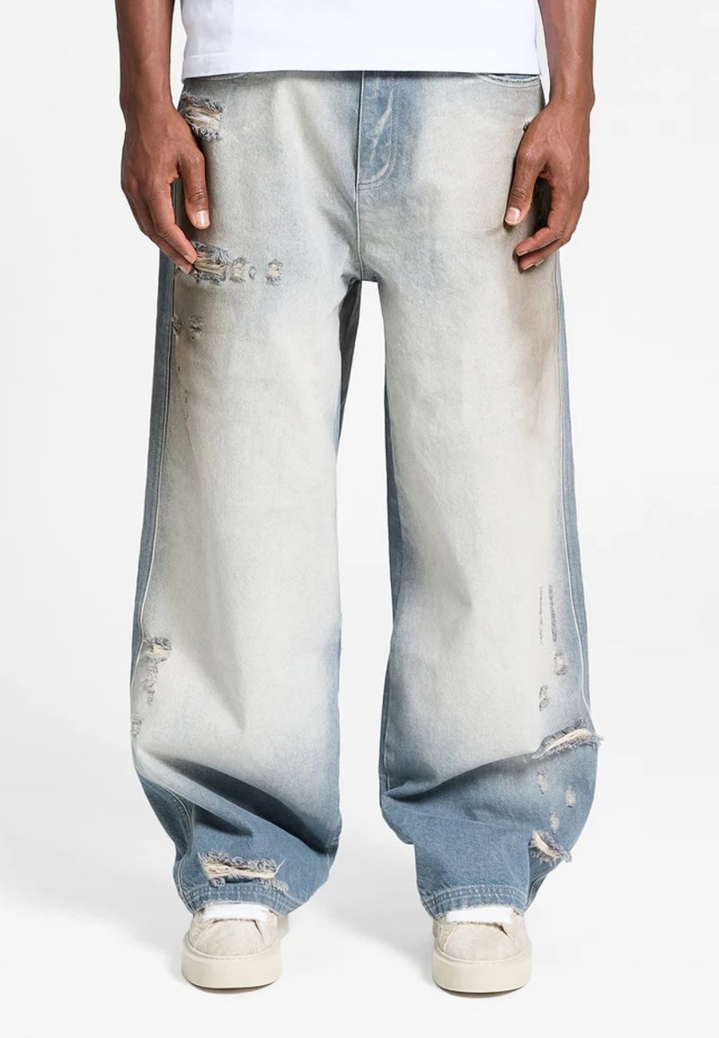 Jeans distressed di colore blu chiaro presentano una vestibilità ampia e svasata con zone sbiadite e orli strappati. Strappi visibili aggiungono texture e dettaglio.
