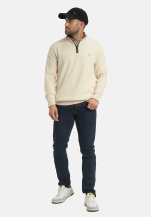 Homme portant un pull beige à col zippé, un jean foncé, des baskets blanches et une casquette noire, debout et regardant vers la gauche sur un fond uni.