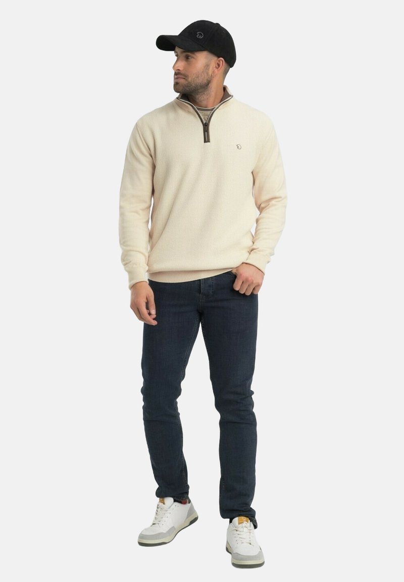 Homme portant un pull beige à col zippé, un jean foncé, des baskets blanches et une casquette noire, debout et regardant vers la gauche sur un fond uni.