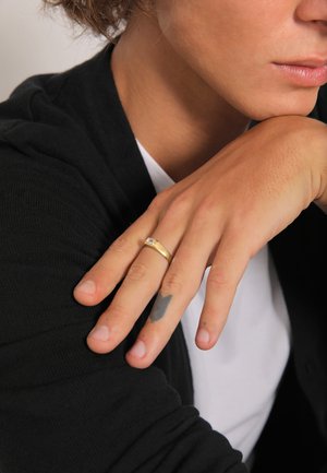 Gold ring with a rectangular clear stone on a hand with light skin and visible tattoos. Background features a black sweater and white shirt.