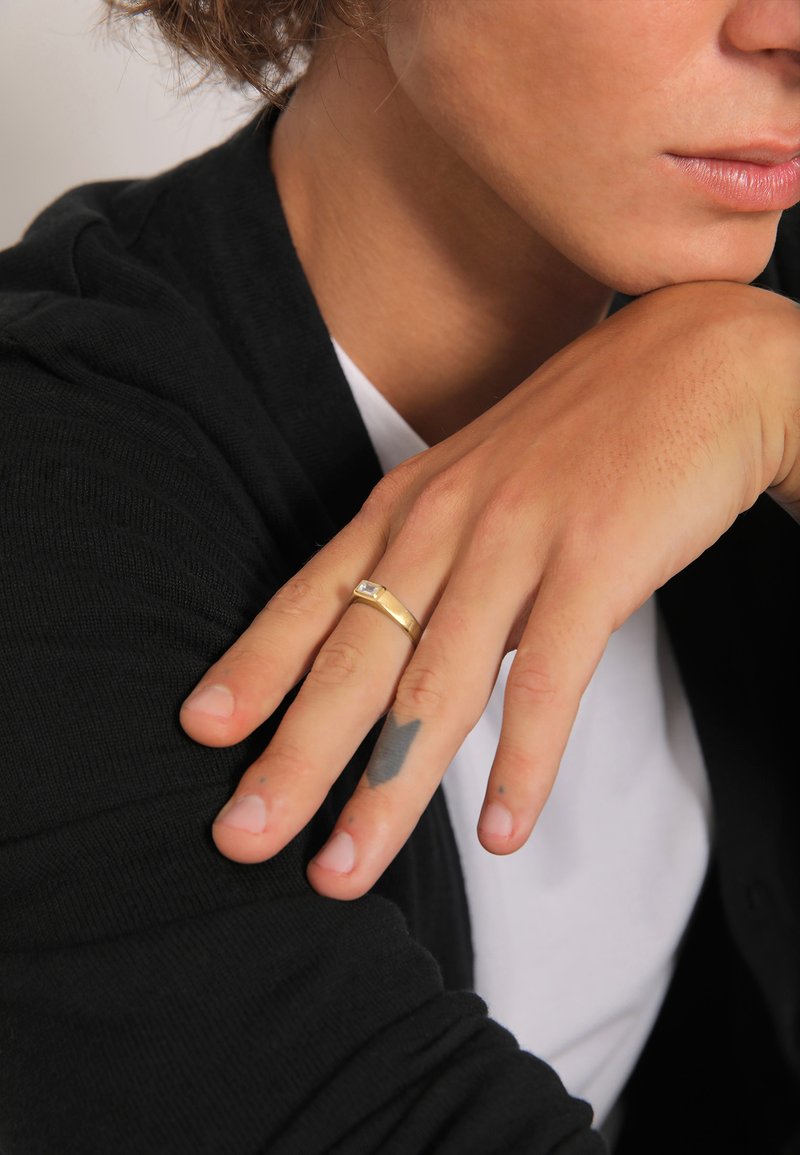 Gouden ring met een rechthoekige heldere steen op een hand met een lichte huid en zichtbare tatoeages. De achtergrond toont een zwarte trui en een witte shirt.
