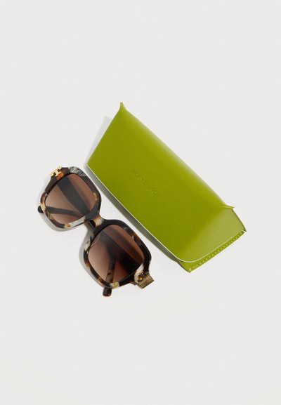 Τετράγωνα γυαλιά ηλίου με καφέ φακούς και σχέδιο χελώνας δίπλα σε κλειστή σκληρή θήκη σε λαχανί χρώμα πάνω σε λευκή επιφάνεια.