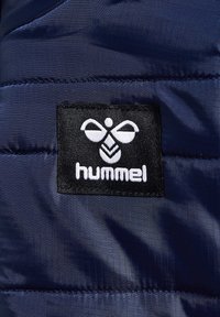 Ein navyfarbener Stoff mit glänzender Textur zeigt ein schwarzes Etikett mit einem Bienenlogo und dem Wort "hummel" in weißer Schrift.
