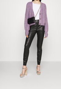 Cardigan tricoté violet clair avec fermeture à boutons, haut blanc, pantalon en cuir noir et sandales à talons argentées avec brides aux chevilles. Sac bandoulière noir.