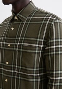 Camicia a quadri verdi con colletto a camicia, motivi bianchi e neri e una tasca sul petto con bottoni marroni chiari. Tessuto in cotone morbido.