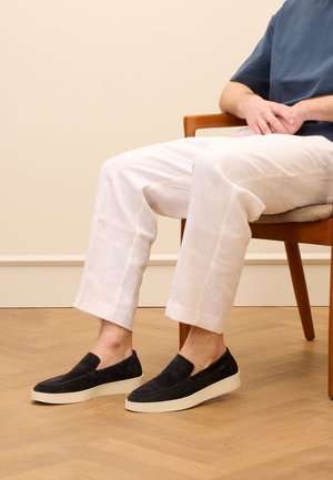 Schwarze Wildleder-Slipper mit einer weißen Gummisohle, kombiniert mit lockeren weißen Leinenhosen, sitzend auf einem Holzstuhl.