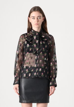 Gjennomsiktig svart bluse med blomsterbroderi i rosa og grønt, med en sløyfe-krage, knappelukking foran, og lange ermer med elastiske mansjetter.