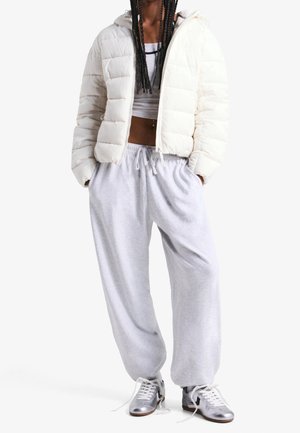 Persoon die een witte cropped pufferjas draagt, lichtgrijze joggingbroek en zilveren sneakers met losgemaakte veters tegen een effen witte achtergrond.
