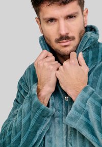 Chaqueta de forro polar color verde azulado con patrón de nervios verticales. Cuello alto, cremallera frontal y textura acogedora. Mangas ajustadas y diseño relajado.