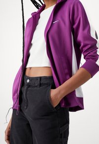 Lila zip-up jacka med vita sidopaneler, gjord av slätt tyg. Bärs över en vit kort topp och kombineras med högmidjade svarta byxor.