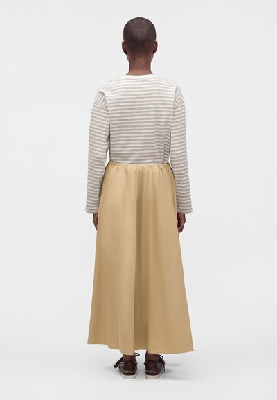 Personne aux cheveux courts portant une longue jupe beige, une chemise rayée à manches longues et des chaussures marron, debout de dos sur un fond uni.