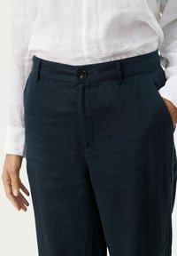 Marineblauwe linnen broek met een relaxte pasvorm, voorzien van een enkele knoopsluiting, zijzakken en een gladde textuur. Gecombineerd met een wit linnen overhemd.