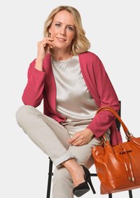 Roze cardigan over een satijnen crème blouse, beige broek en zwarte hakken; vasthoudend een oranje leren handtas met trekkoorddetail.